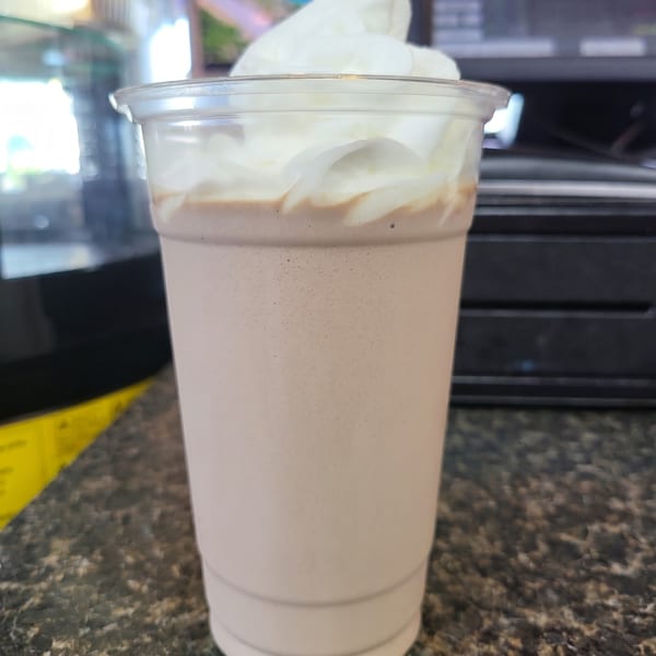 A chocolate milkshake with whipped cream on top, in a clear plastic cup on a dark counter.
