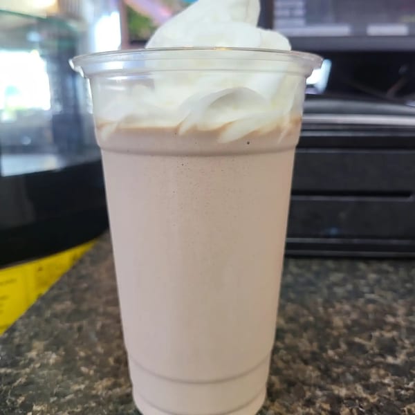 A chocolate milkshake with whipped cream in a clear plastic cup on a dark speckled counter.