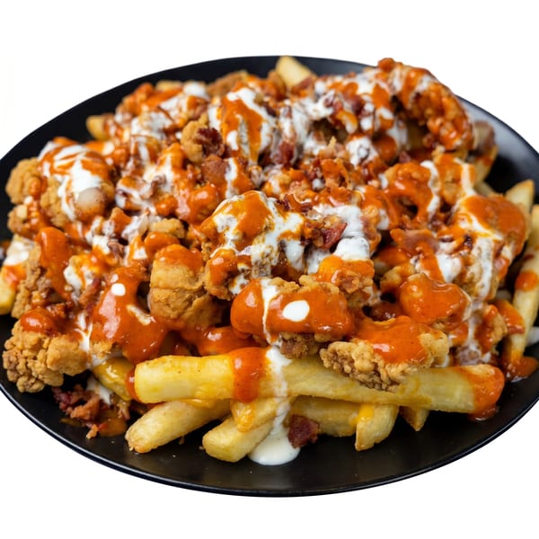 Crispy chicken bites, bacon, and fries smothered in buffalo sauce and ranch dressing on a black plate.